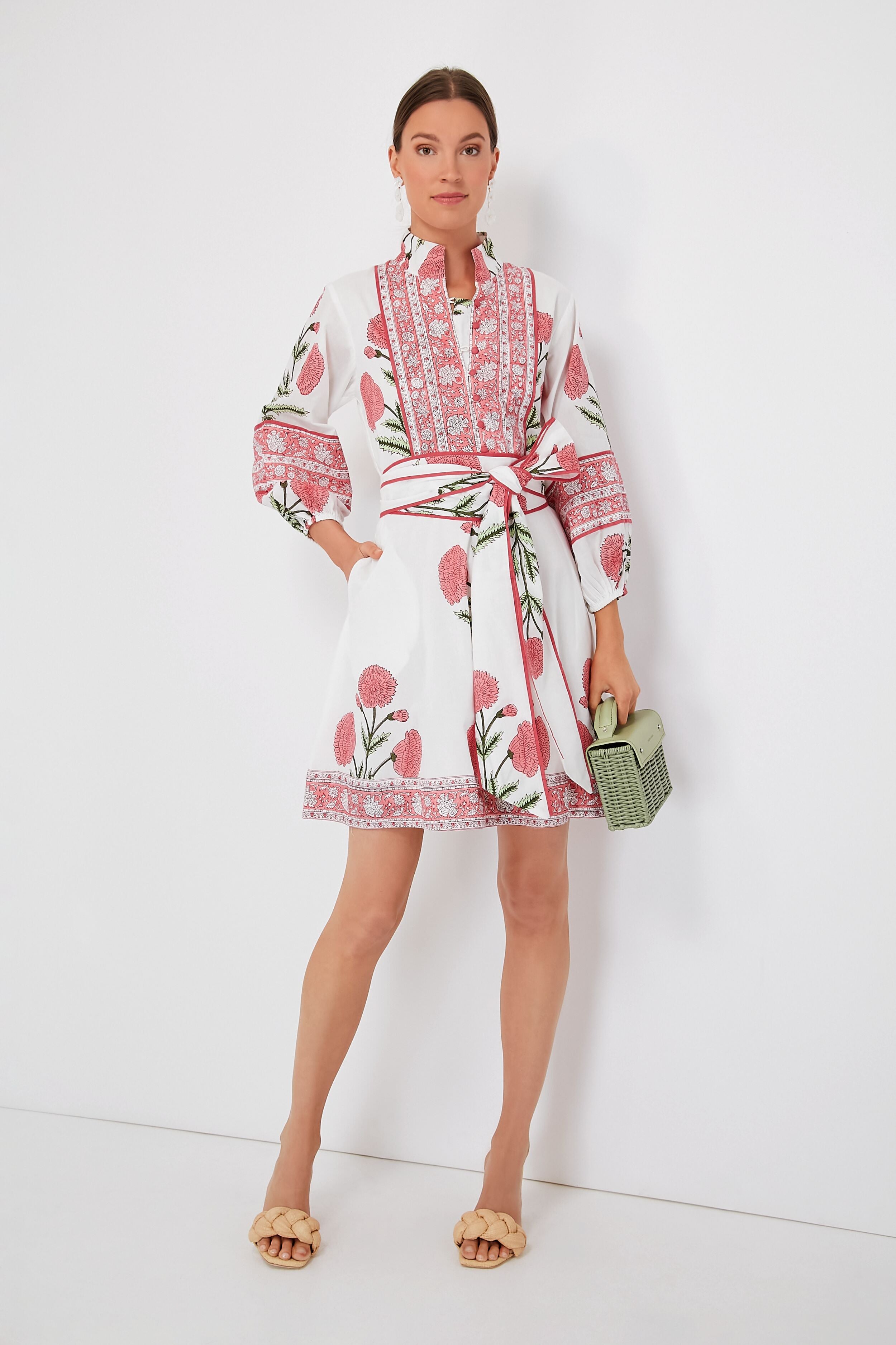 A woman wears the Sue Sartor Rosy Marigold Flounce Shorty Dress, featuring pink floral patterns and a matching waist tie, paired with a green woven handbag and beige sandals—crafted from sustainable fabrics for a modern look.