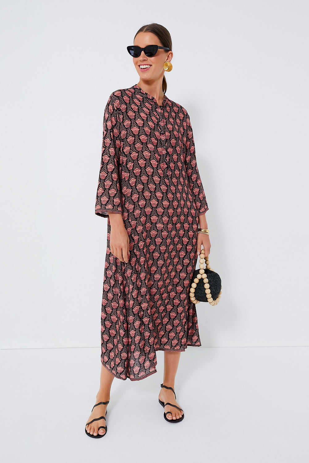 A woman in a Natalie Martin batik print, long-sleeved, ankle-length dress pairs it with black sandals, sunglasses, large earrings, and carries a black handbag accented with white beads.