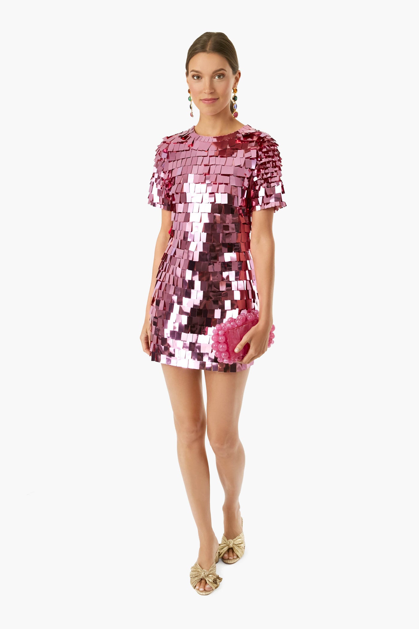 A woman wears a SIMKHAI sequin mini dress with square reflective embellishments, gold open-toe heels, colorful earrings, and carries a pink clutch.