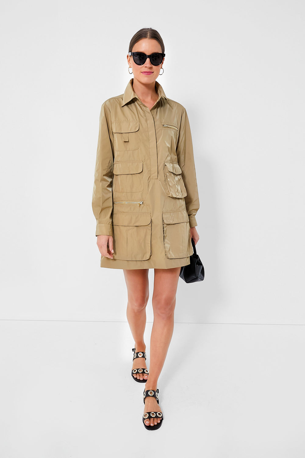 A woman sports a Cynthia Rowley safari-chic khaki backpack dress, paired with black sunglasses, gold-accented black sandals, and a small black handbag.