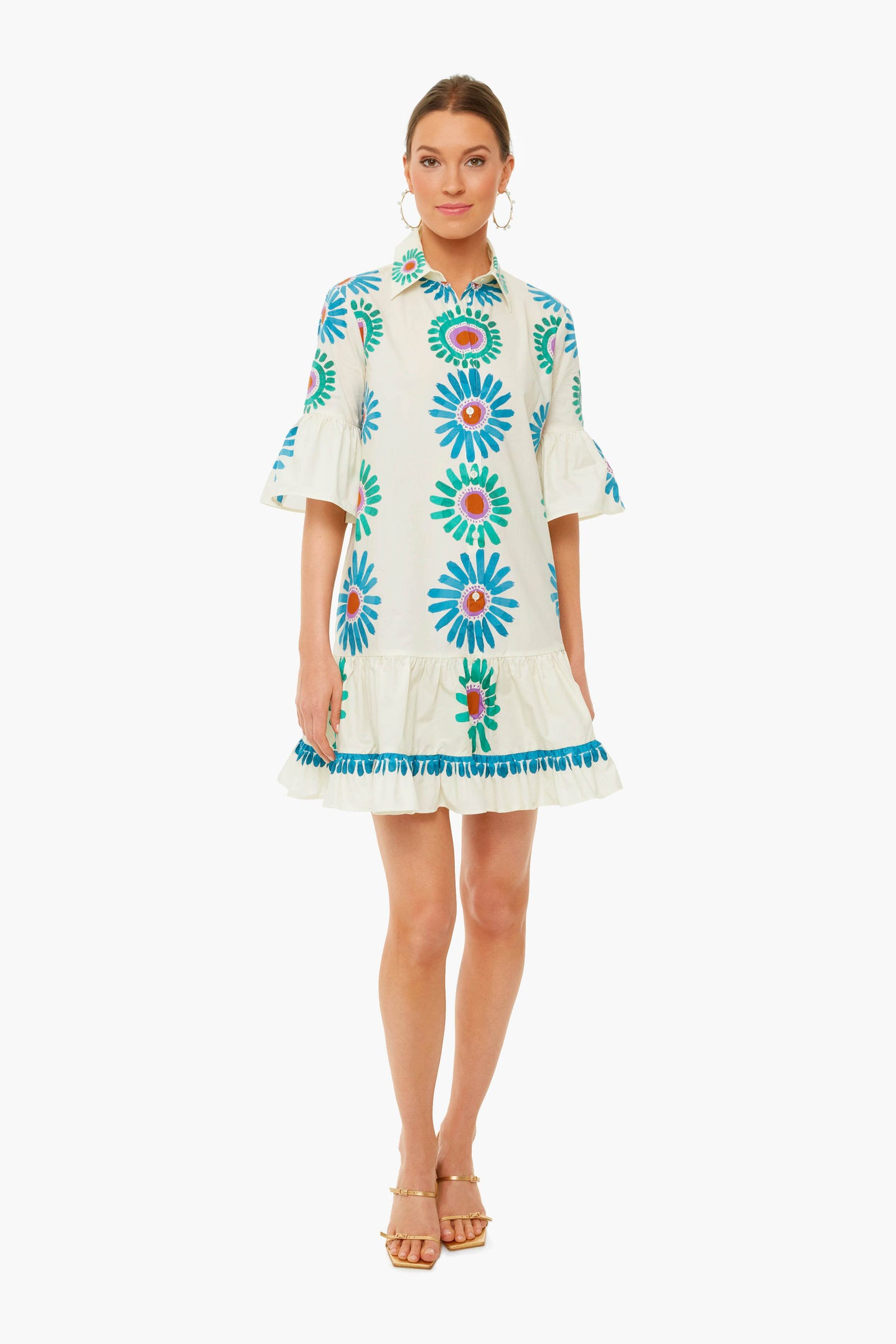 A woman wears a La DoubleJ knee-length white cotton poplin dress with blue and orange floral patterns, flared sleeves, a ruffled hem, and gold sandals.