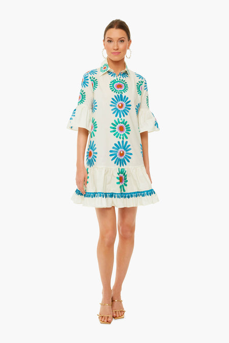 A woman wears a La DoubleJ knee-length white cotton poplin dress with blue and orange floral patterns, flared sleeves, a ruffled hem, and gold sandals.