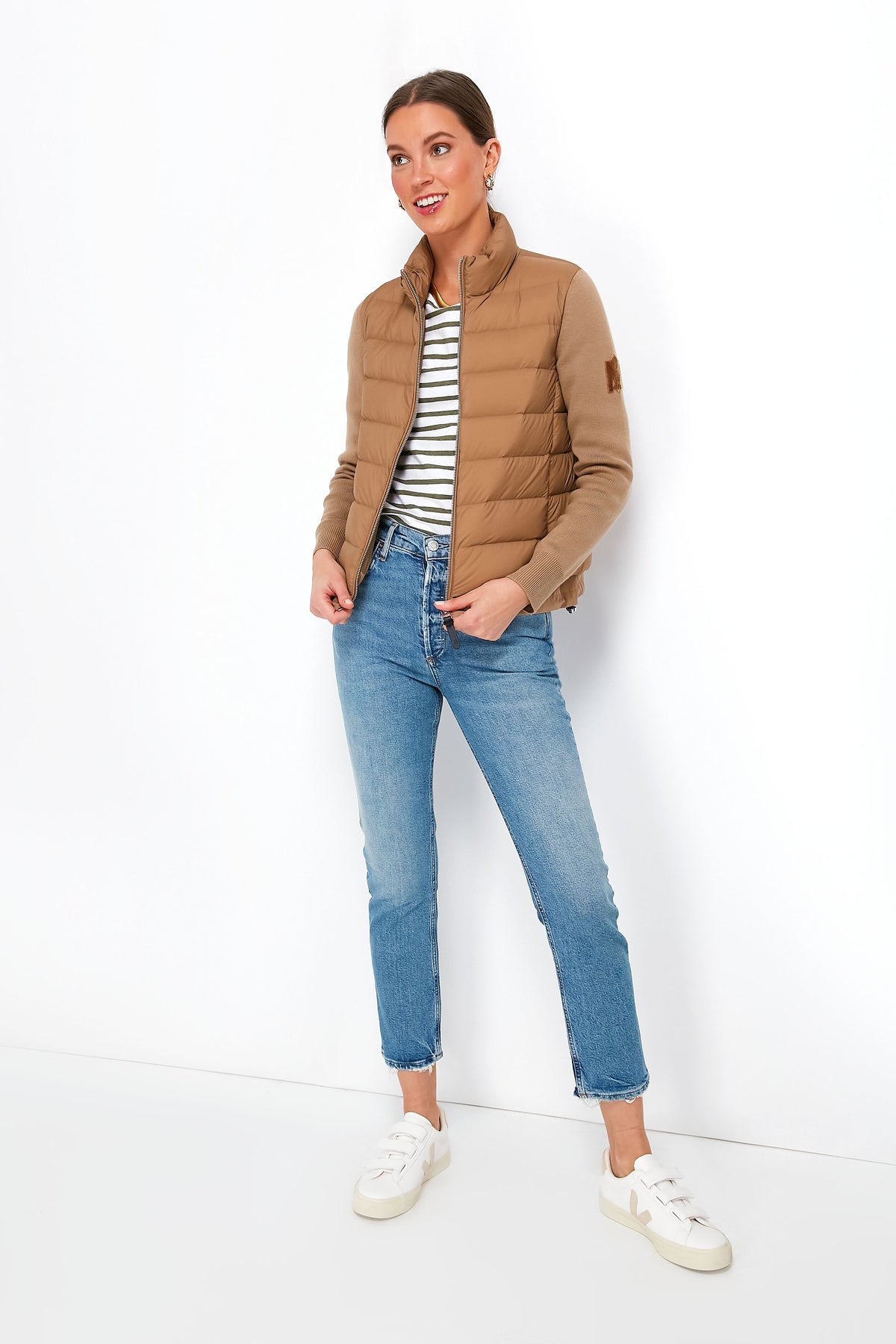 A woman in a tan MACKAGE jacket, striped shirt, blue jeans, and white sneakers stands with her hands in her jacket pockets.