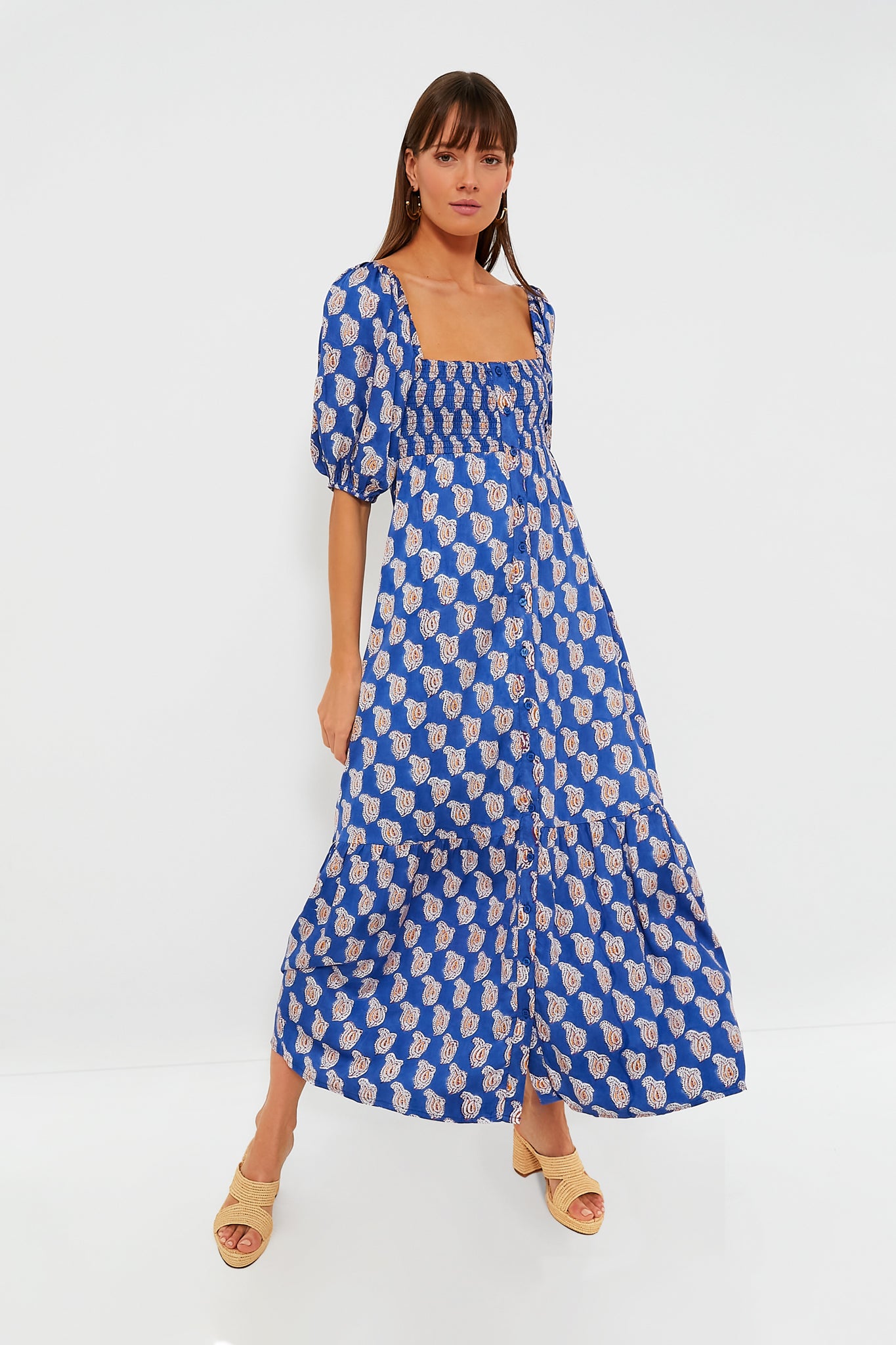 A woman wears a Love Tomas by Emerson Fry hand block printed blue maxi dress with a beige leaf pattern, puff sleeves, and square neckline, paired with beige sandals.