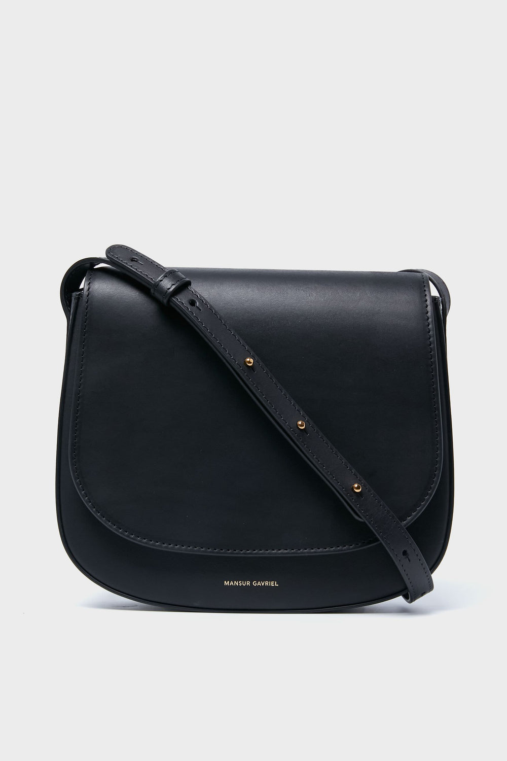 A Black Classic Crossbody bag by Mansur Gavriel, made from vegetable tanned leather with a flap closure, adjustable strap, and the brand name in gold lettering on the front.