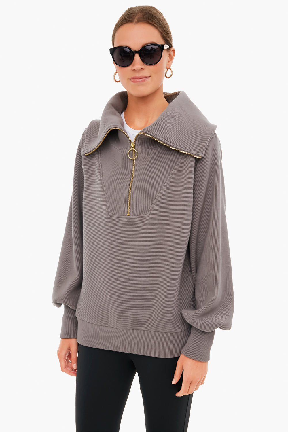 A woman in black sunglasses, gold hoop earrings, and a Varley half zip pullover with a wide collar and balloon sleeves stands confidently, highlighting Varley's signature blend of performance and style.