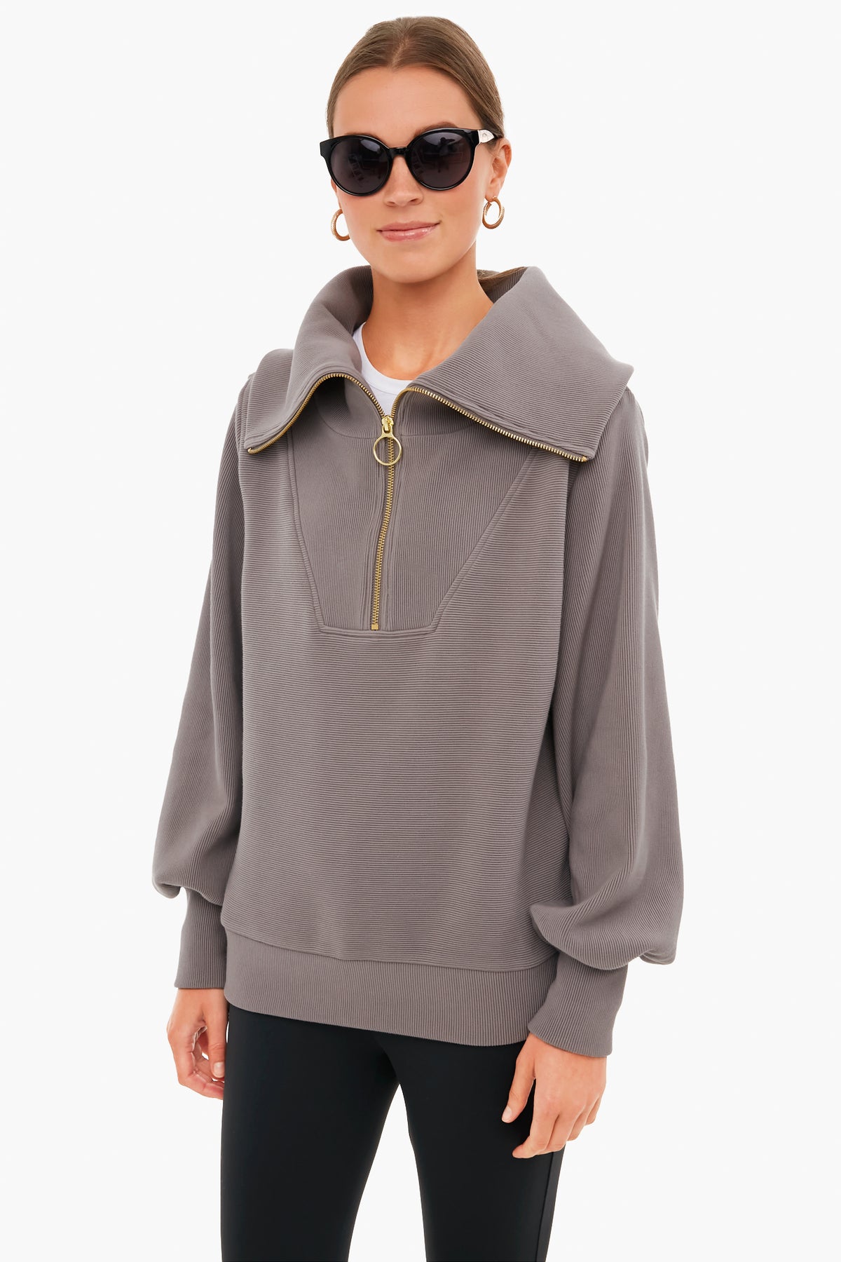 A woman in black sunglasses, gold hoop earrings, and a Varley half zip pullover with a wide collar and balloon sleeves stands confidently, highlighting Varley's signature blend of performance and style.