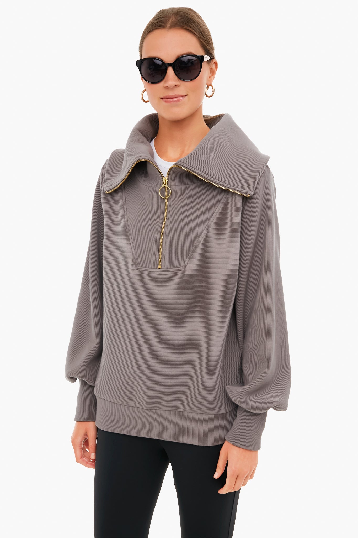A woman in black sunglasses, gold hoop earrings, and a Varley half zip pullover with a wide collar and balloon sleeves stands confidently, highlighting Varley's signature blend of performance and style.