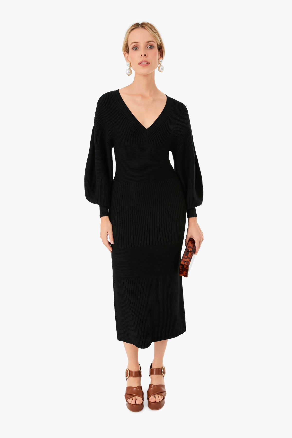 A woman wears a STAUD Black Carnation sweater midi dress featuring long sleeves and a V-neck, paired with brown platform sandals, statement earrings, and holding a small brown clutch.