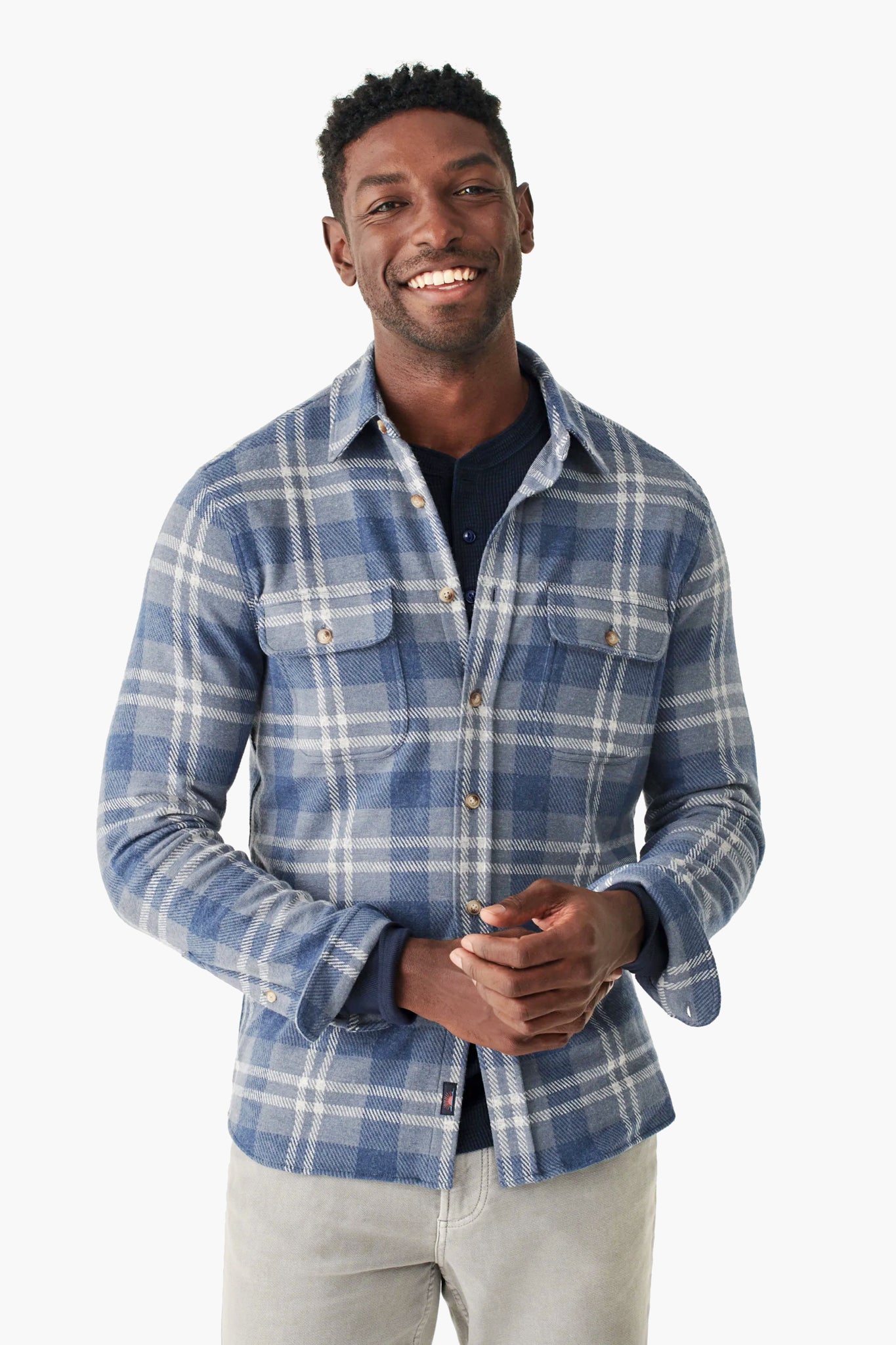 A man in a Faherty blue and white vintage plaid sweater shirt over a dark top and light gray pants stands smiling, showcasing effortless fall layering.