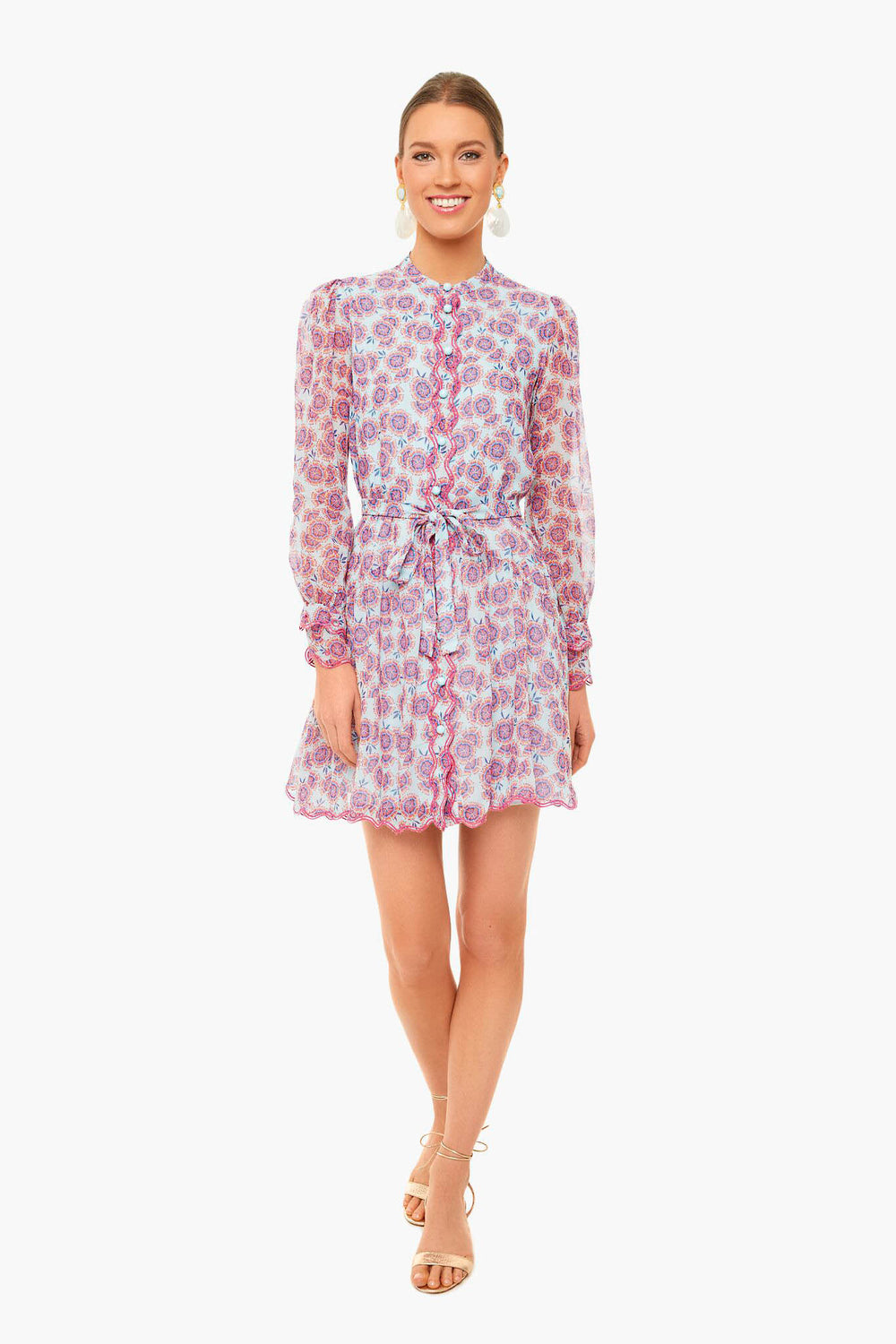 Woman standing and smiling in a Saloni Tilly Dress—long-sleeve, button-up, floral print with a tied waist—paired with beige sandals.