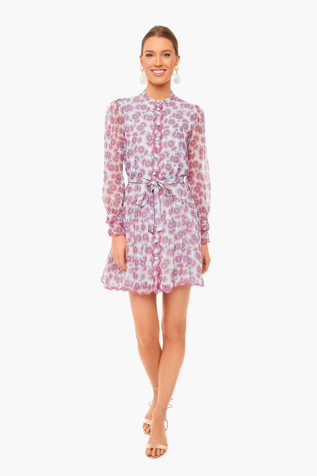 Woman standing and smiling in a Saloni Tilly Dress—long-sleeve, button-up, floral print with a tied waist—paired with beige sandals.