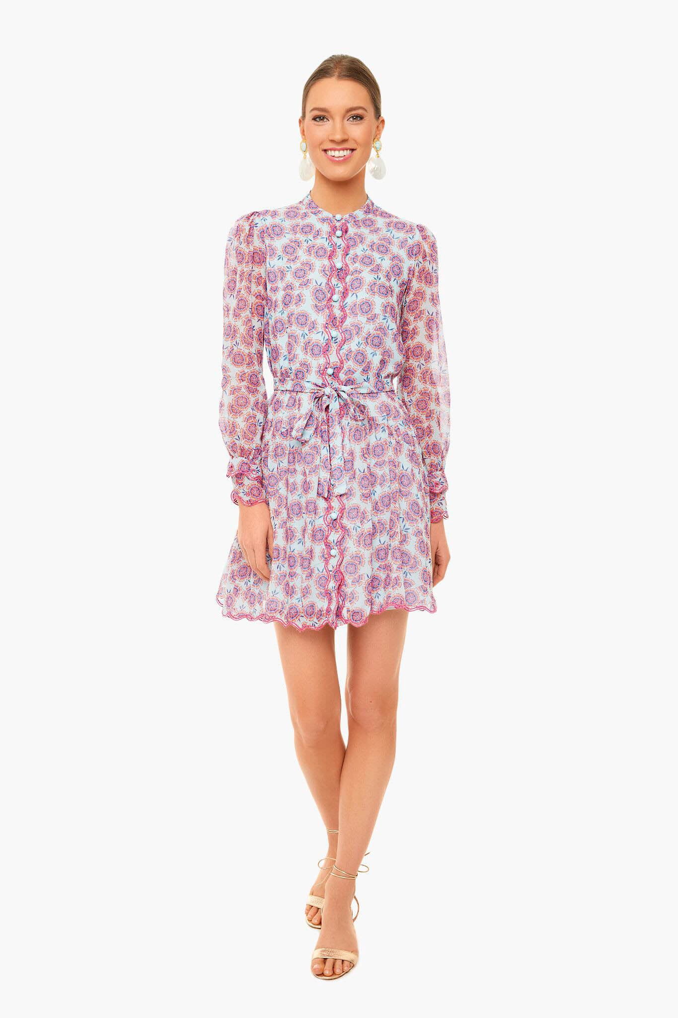 Woman standing and smiling in a Saloni Tilly Dress—long-sleeve, button-up, floral print with a tied waist—paired with beige sandals.