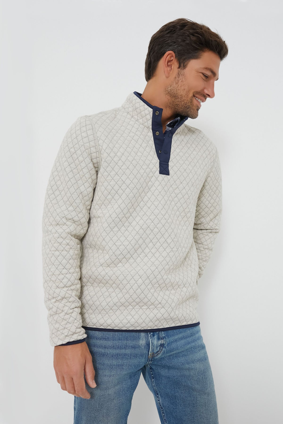 A man wearing a Marine Layer light-colored, quilted, sustainable pullover with a dark buttoned collar and blue jeans stands looking to his right.