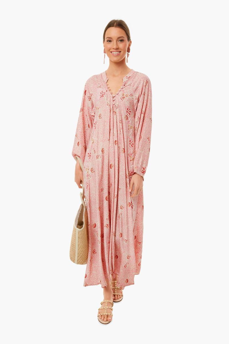 A woman wears a Natalie Martin long, loose-fitting pink floral silk maxi dress with long sleeves, paired with a woven tote bag and flat sandals.