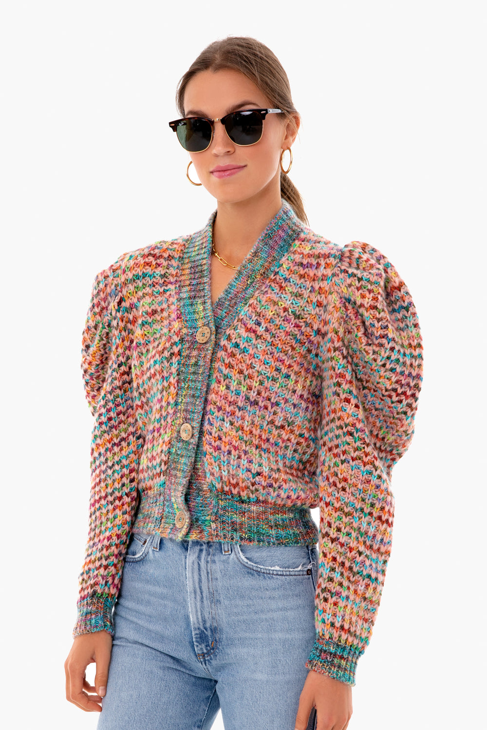 A woman stands confidently in black sunglasses, gold hoop earrings, blue jeans, and a colorful Ulla Johnson merino wool cardigan featuring voluminous sleeves.