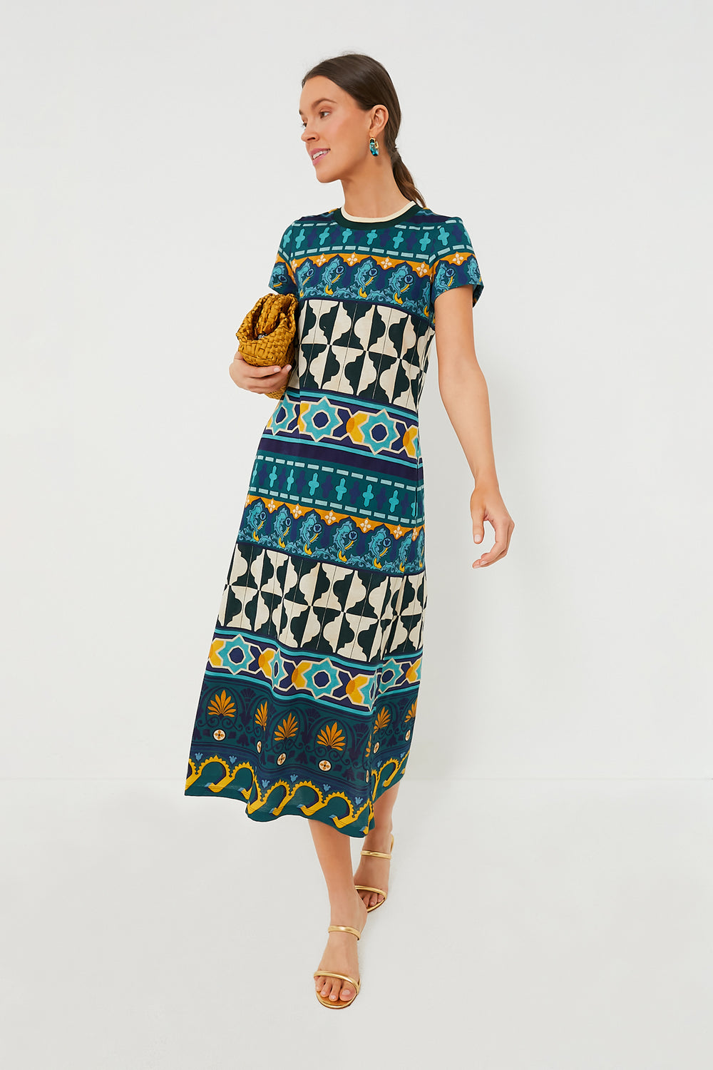A woman in a La DoubleJ patterned short-sleeve cotton jersey dress with geometric and floral designs, holding a woven clutch, paired with sandals and earrings.