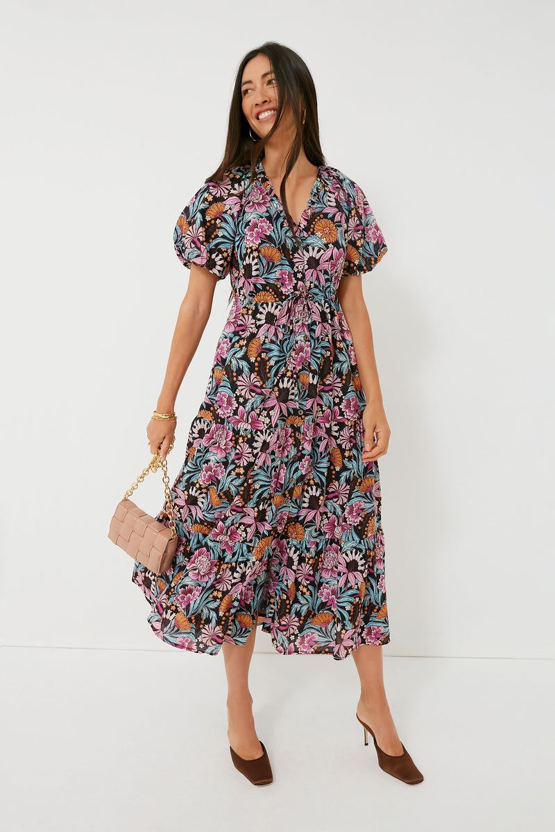 A woman in a colorful Banjanan Black Beauty Poppy Dress made from organic cotton voile stands holding a pink quilted purse and wearing brown heels.