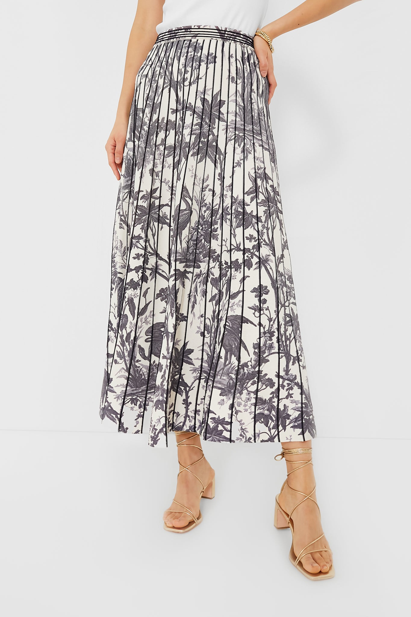 A person wears a Cara Cara white midi skirt with black botanical prints, pleats, and tan strappy sandals.