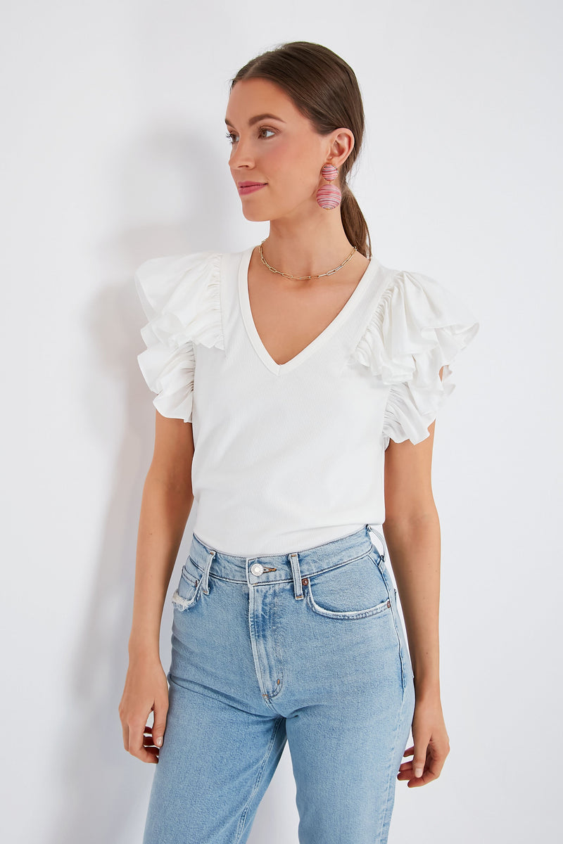 A woman wearing a Pomander Place white flutter sleeve top and light blue jeans stands, looking to the side.