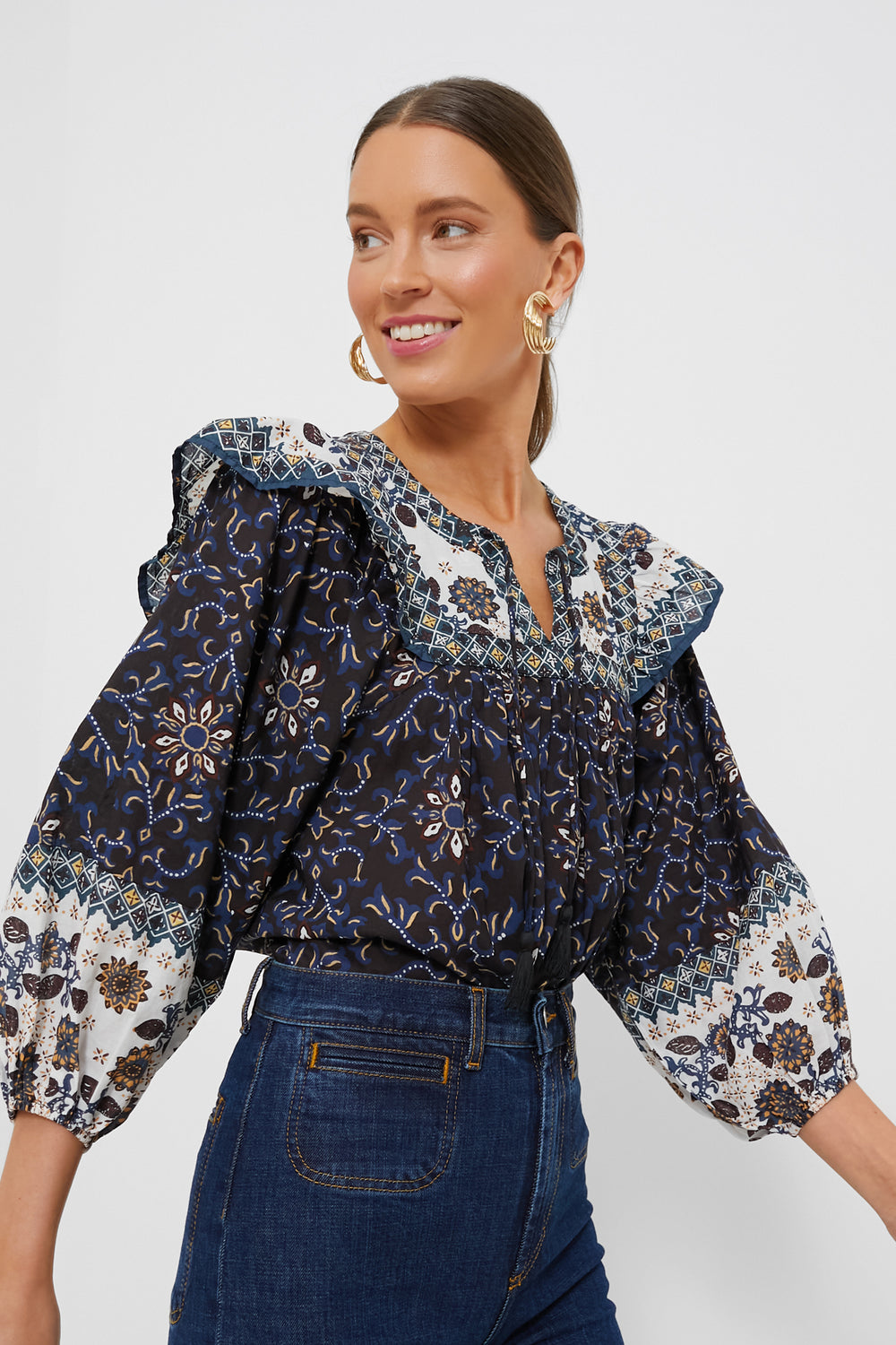 A woman models a Cleobella navy and white organic cotton folk-inspired blouse with ruffled shoulders, paired with dark blue high-waisted jeans—both ethically handmade garments.
