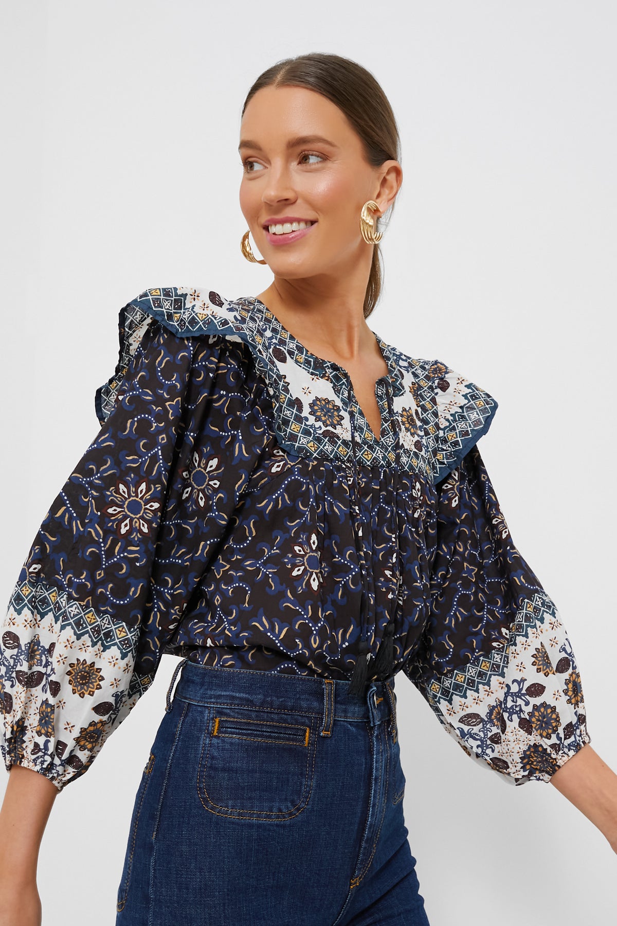 A woman models a Cleobella navy and white organic cotton folk-inspired blouse with ruffled shoulders, paired with dark blue high-waisted jeans—both ethically handmade garments.