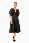A woman stands wearing a Ulla Johnson black cotton poplin midi dress with puff sleeves and a V-neckline, paired with black open-toed shoes.