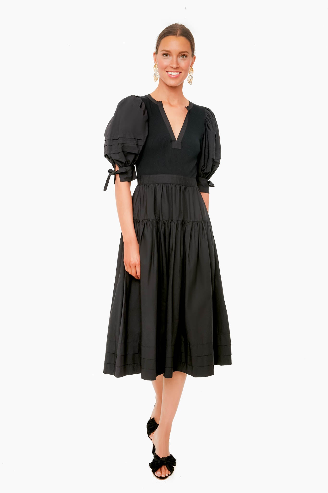 A woman stands wearing a Ulla Johnson black cotton poplin midi dress with puff sleeves and a V-neckline, paired with black open-toed shoes.