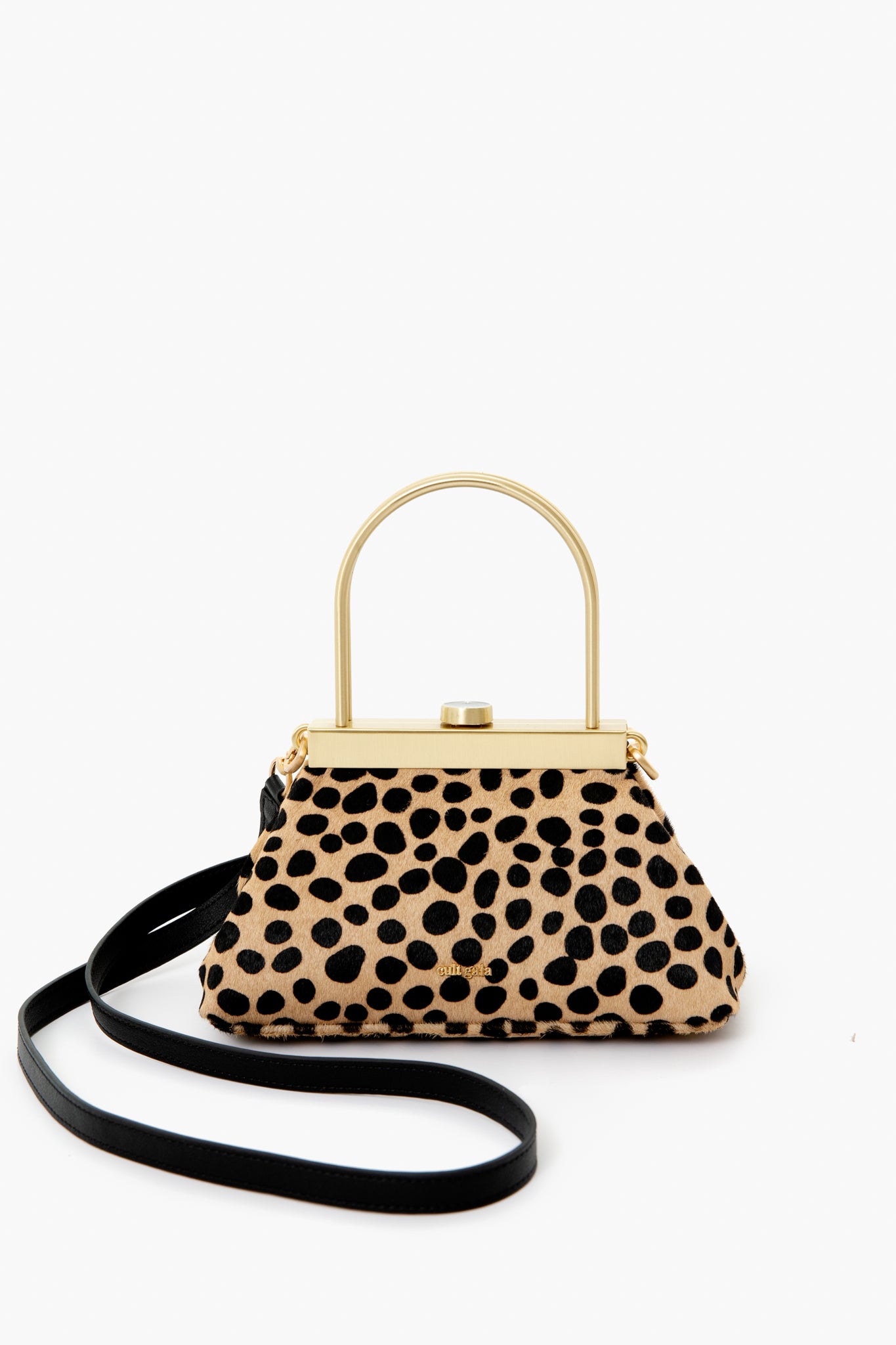 The Cult Gaia bag features a sculptural design with a gold metal top handle, black shoulder strap, and beige fabric adorned with black irregular dots, making it a unique accessory for any outfit.