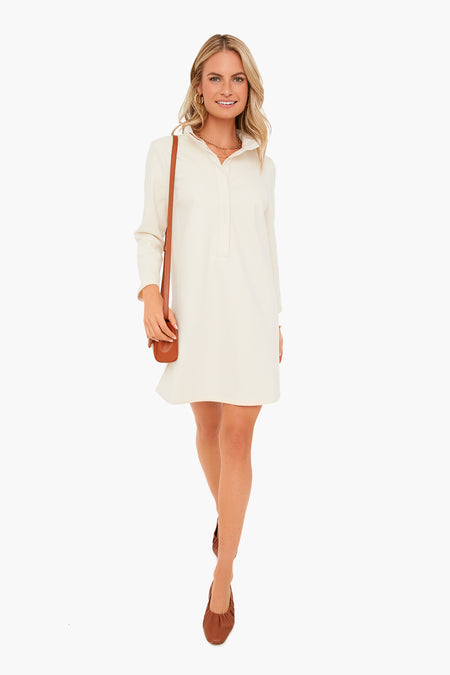 A woman smiles while standing in a Tuckernuck long-sleeve stretch knit white collared dress, paired with brown flats and a matching brown shoulder bag.