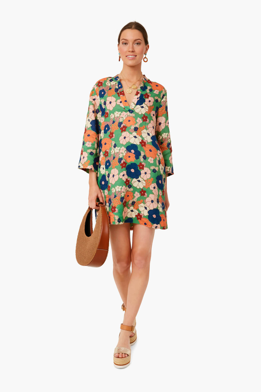 A woman in a colorful floral shift dress by TROVATA Birds of Paradis and wedge sandals holds a round brown bag, smiling brightly.