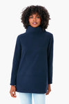 A woman in a Dudley Stephens navy blue turtleneck sweater, made from sustainable recycled yarn, and light blue jeans stands facing forward, showcasing the cozy stand collar.
