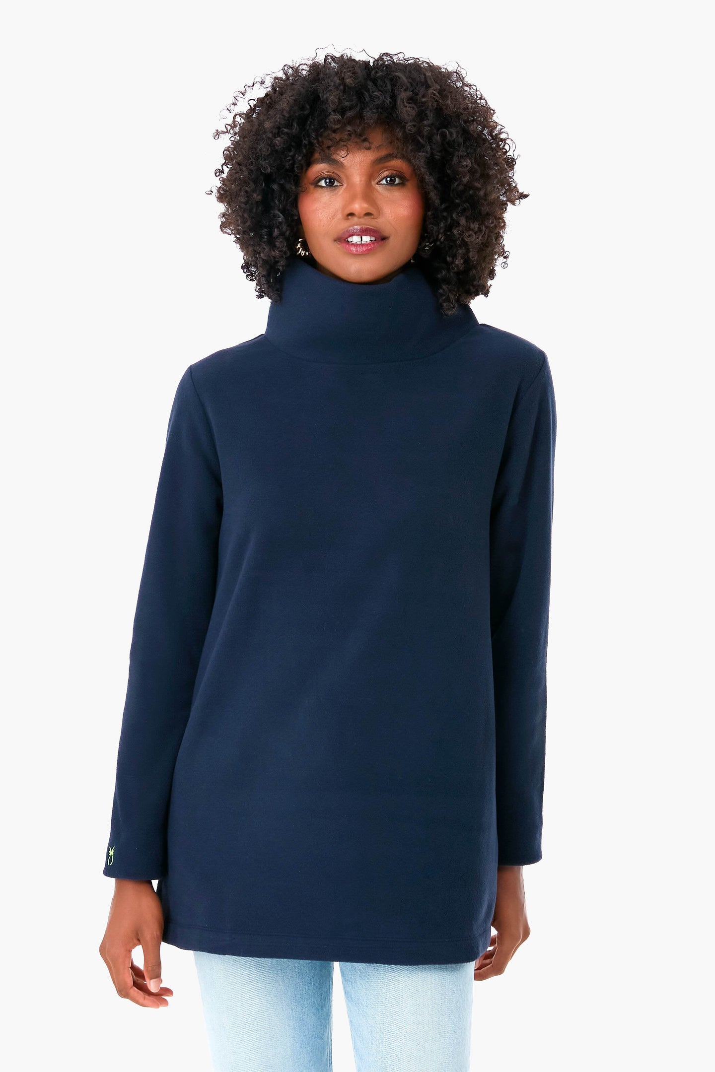 A woman in a Dudley Stephens navy blue turtleneck sweater, made from sustainable recycled yarn, and light blue jeans stands facing forward, showcasing the cozy stand collar.
