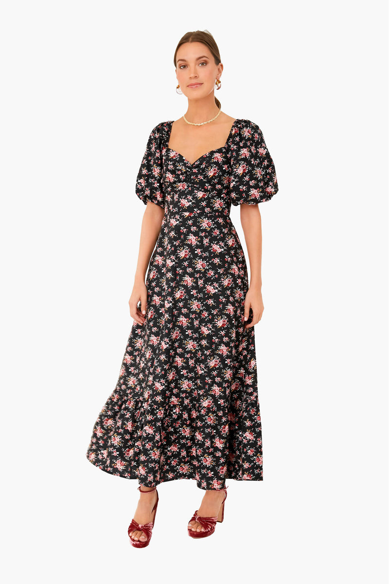 A woman exudes elegance in a Hyacinth House black floral midi dress with puff sleeves and a sweetheart neckline, styled with a pearl necklace and red open-toe heels.
