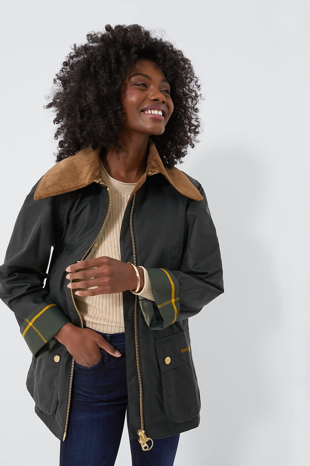 A woman wearing a Barbour jacket with a brown corduroy collar, cream sweater, and dark jeans stands smiling and looking to the side.