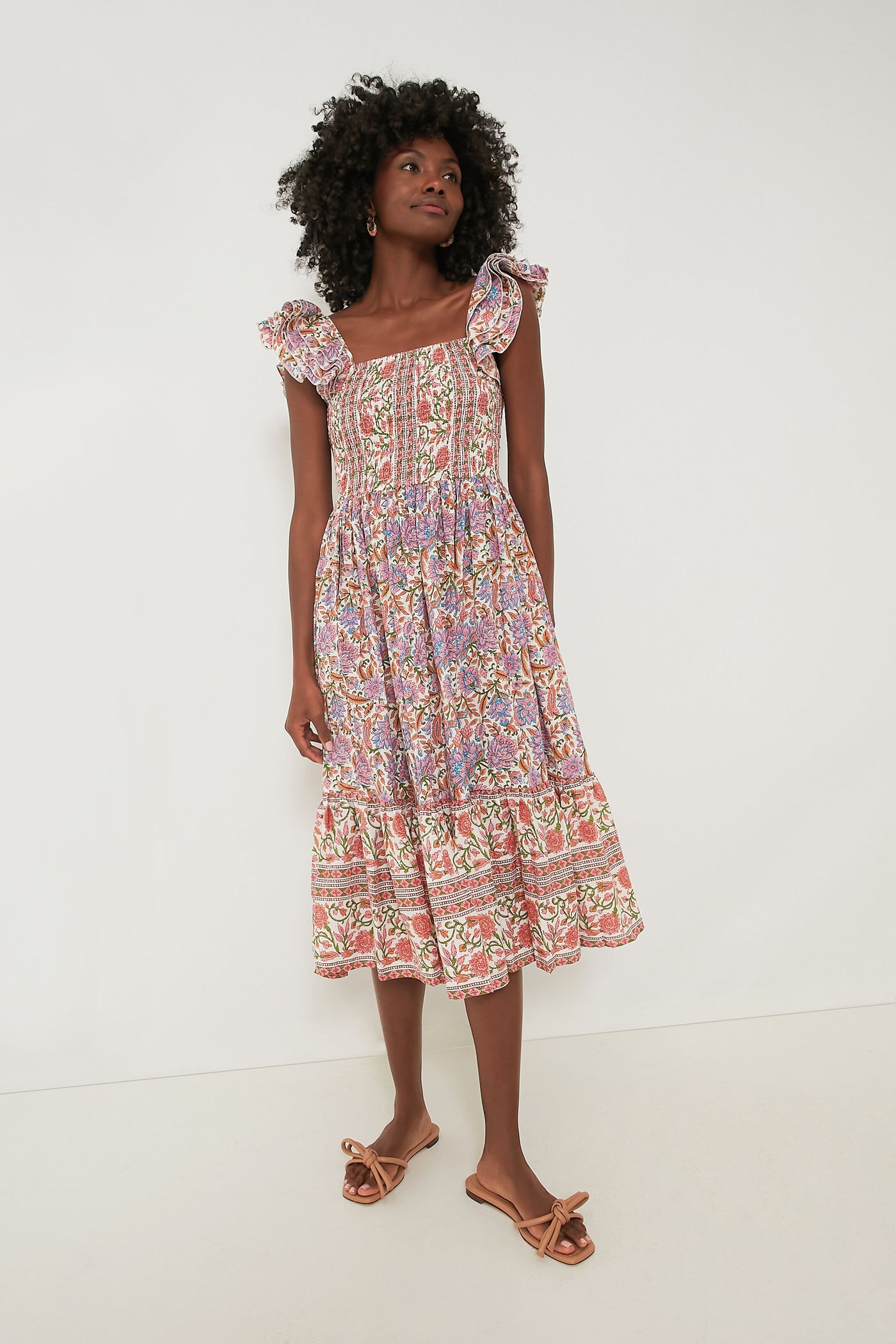 A woman wears the Mille Avignon Floral Olympia Dress, a sleeveless, floral midi dress with ruffled straps and a smocked bodice, paired with bow sandals.