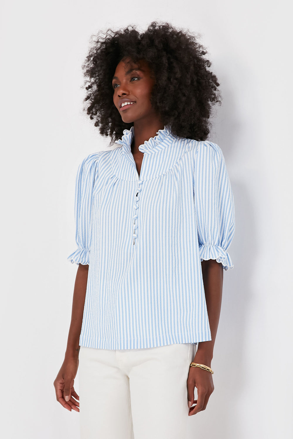 A woman wears a light blue and white vertical striped Pomander Place Selena Blouse with a ruffled collar and elbow-length puff sleeves in airy seersucker fabric, paired with white pants.