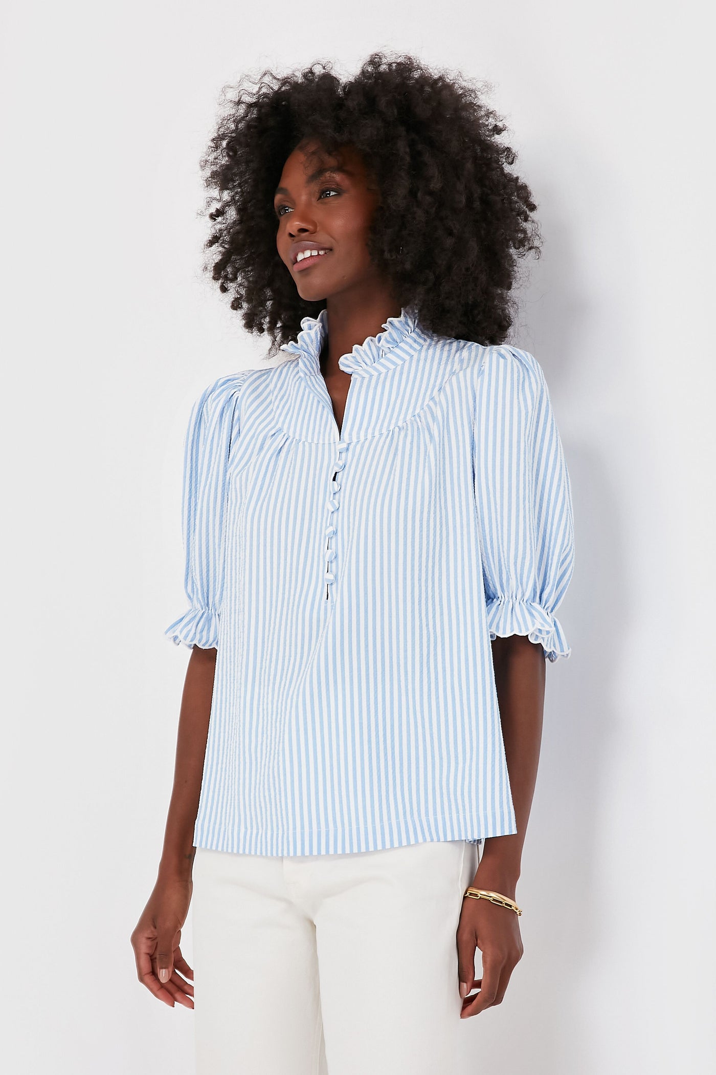 A woman wears a light blue and white vertical striped Pomander Place Selena Blouse with a ruffled collar and elbow-length puff sleeves in airy seersucker fabric, paired with white pants.