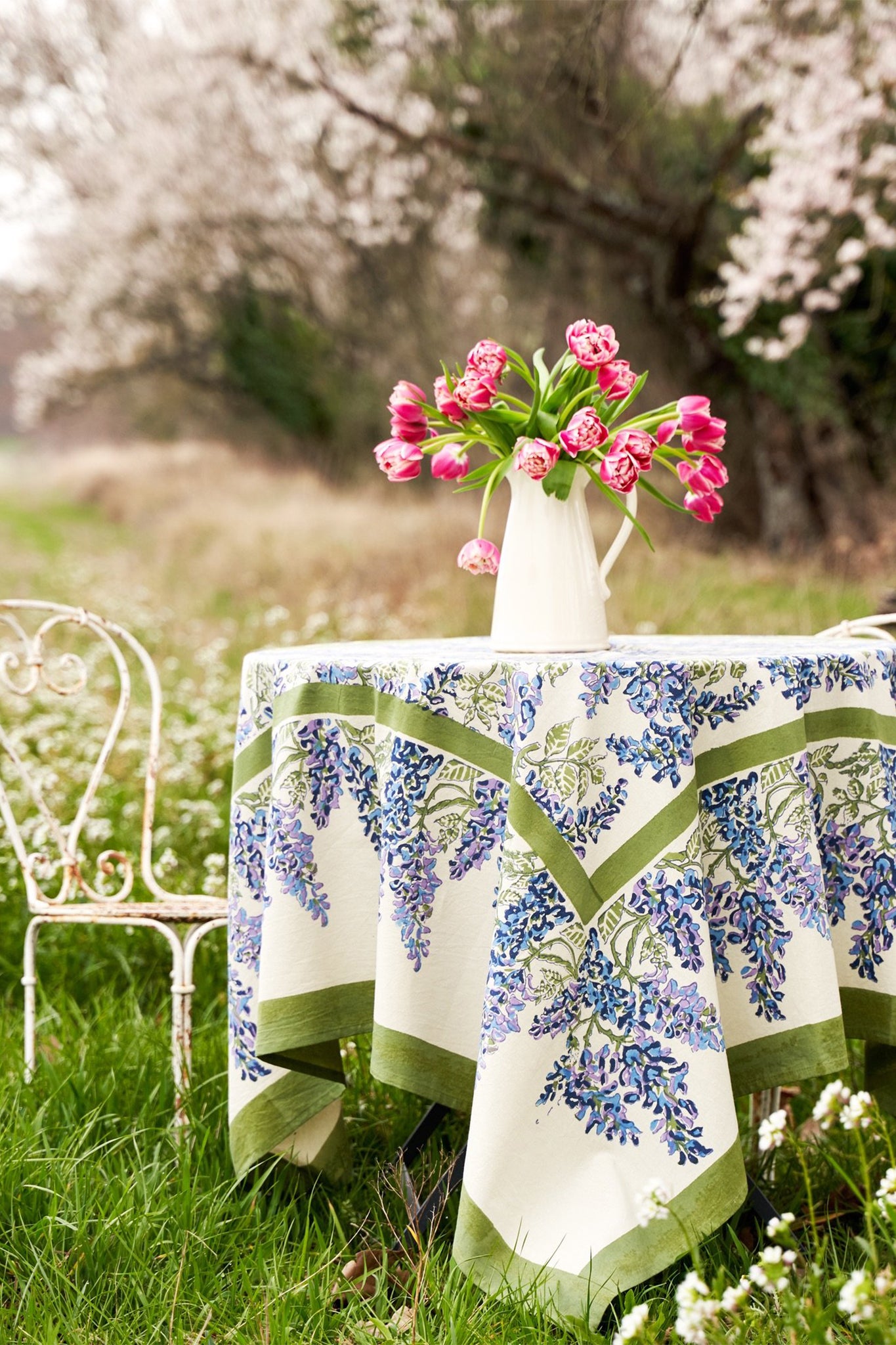 A round table with a Couleur Nature Paris floral table linen and a white pitcher of pink tulips sits on grass next to a white metal chair, surrounded by blooming trees.