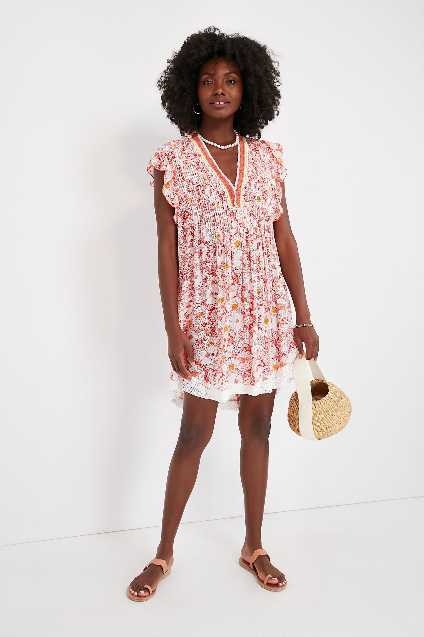 A woman wears the boho-chic Sasha Dress by Poupette St Barth, styled with ruffle sleeves and a floral print. She pairs it with sandals, layered necklaces, and a round straw bag for a breezy summer look.