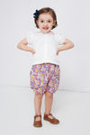 A young child dressed in a Rachel Riley daisy embroidered blouse, floral shorts, brown shoes, and a dark hair bow.