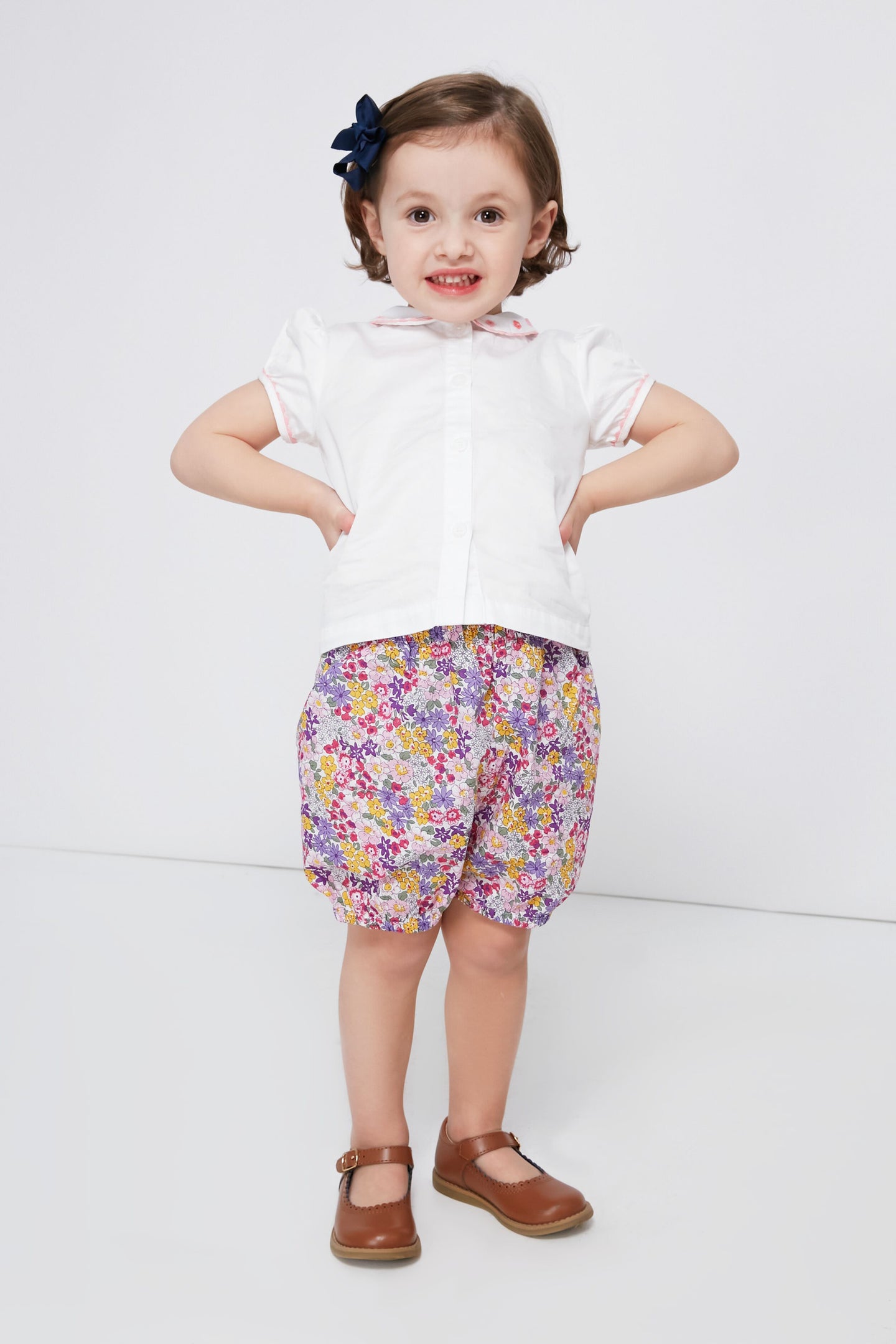 A young child dressed in a Rachel Riley daisy embroidered blouse, floral shorts, brown shoes, and a dark hair bow.