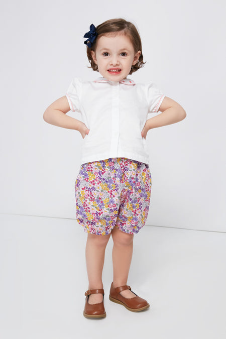 A young child dressed in a Rachel Riley daisy embroidered blouse, floral shorts, brown shoes, and a dark hair bow.