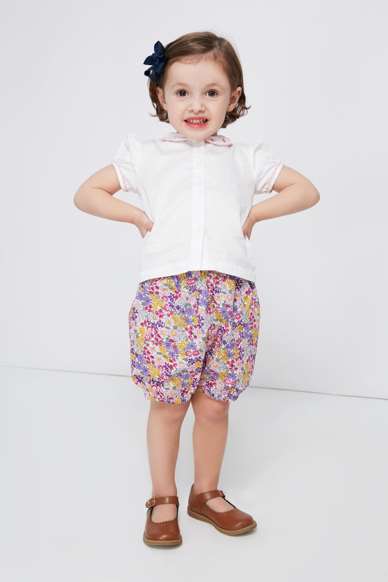 A young child dressed in a Rachel Riley daisy embroidered blouse, floral shorts, brown shoes, and a dark hair bow.