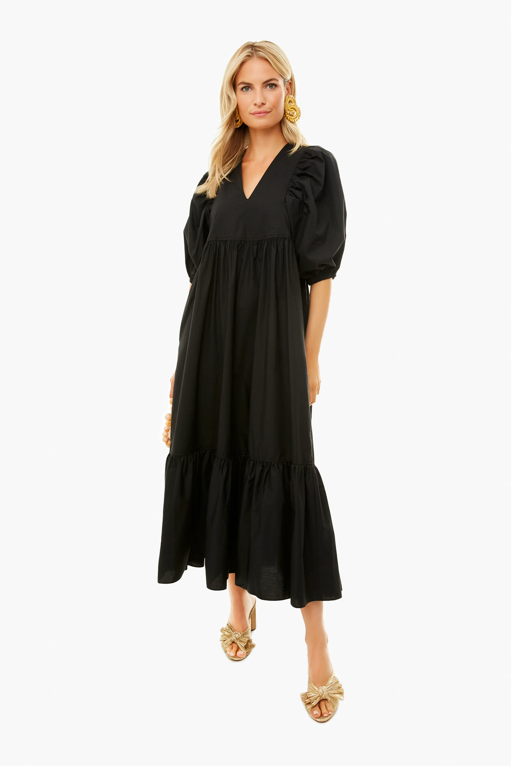 Woman wearing a black Hunter Bell Pretty Palmer Dress with puff sleeves, V-neckline, and tiered ruffle hem, styled with gold statement earrings and gold slip-on sandals.