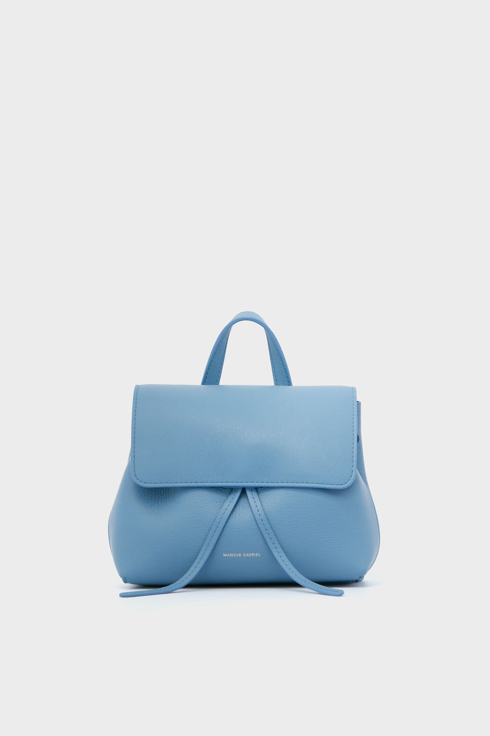 The Mansur Gavriel mini lady bag is crafted from Italian leather in light blue, featuring a top handle, front flap closure, and subtle gold text at the base.