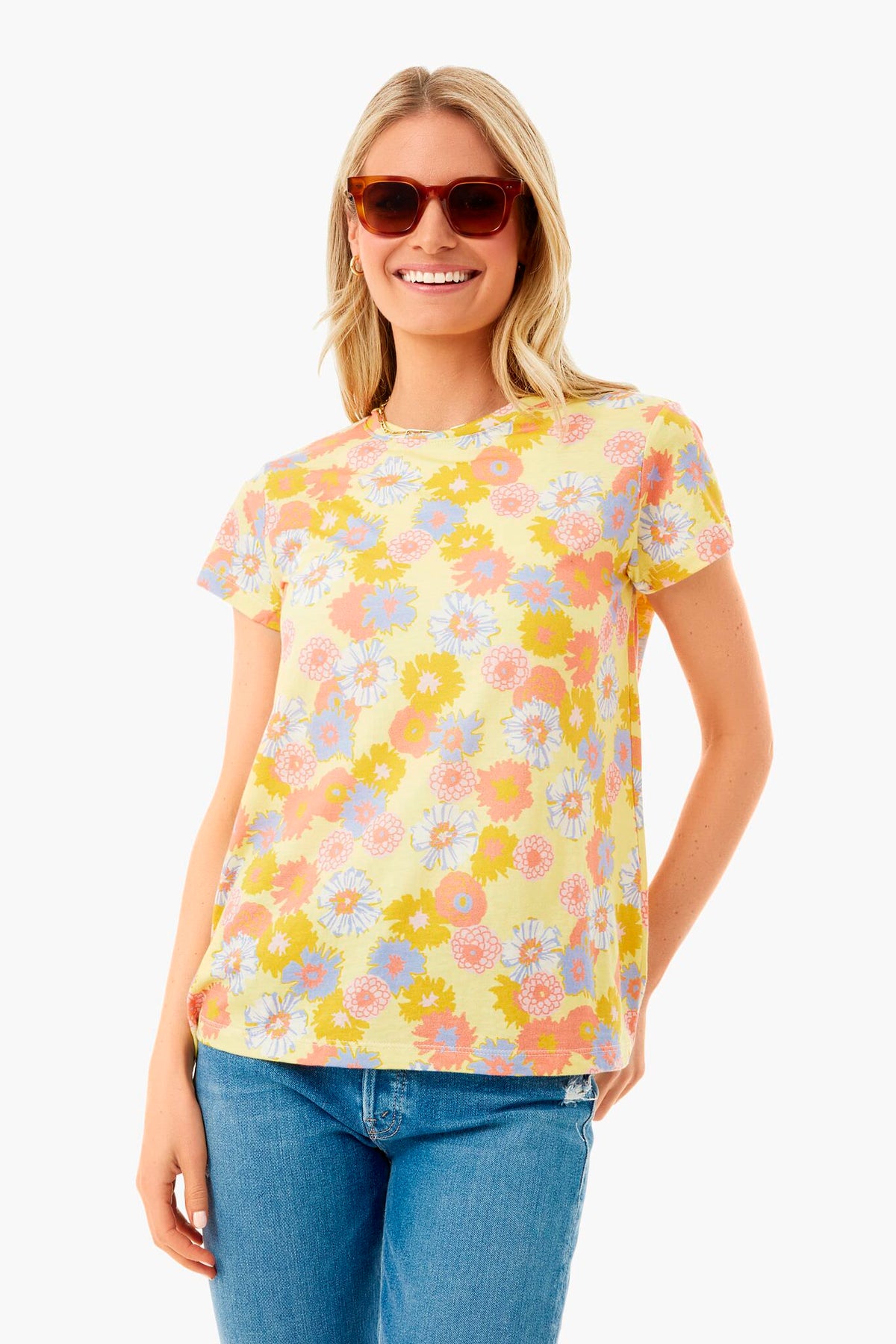A woman wearing sunglasses, a Marine Layer yellow vintage floral crew, and blue jeans smiles in a brightly lit space.
