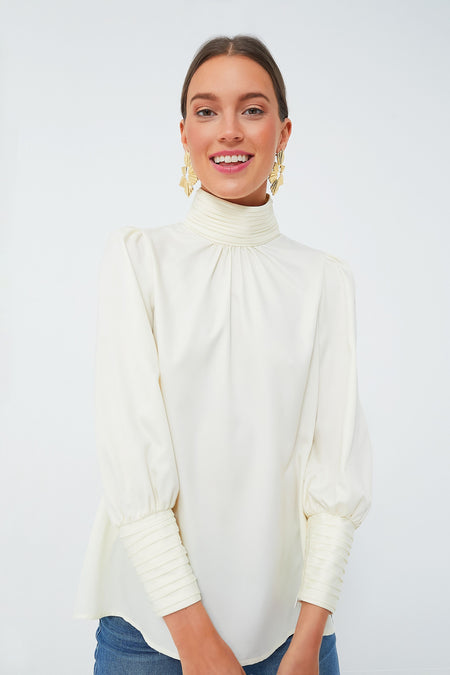 A woman wears a long-sleeve, high-neck cream Tuckernuck Faven Blouse with pleated cuffs and gold statement earrings, styled with a modern twist.