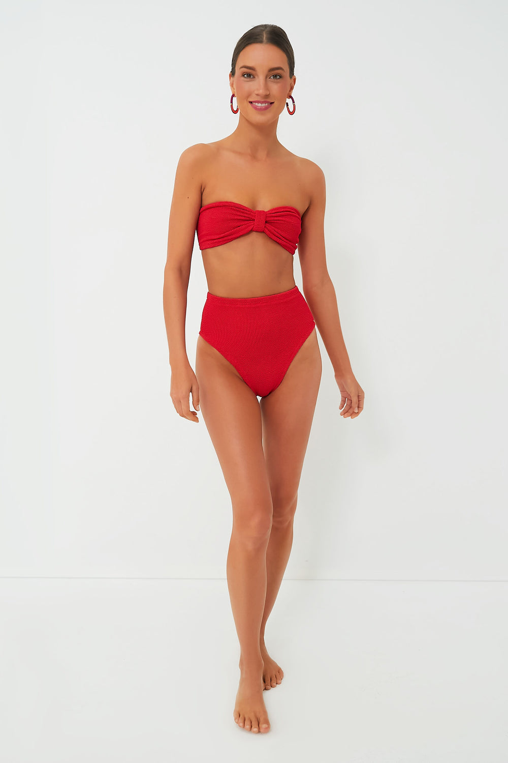 A woman stands barefoot in a Hunza G red crinkle-stretch bikini featuring a bow detail and retro high-waisted bottoms, paired with red hoop earrings.
