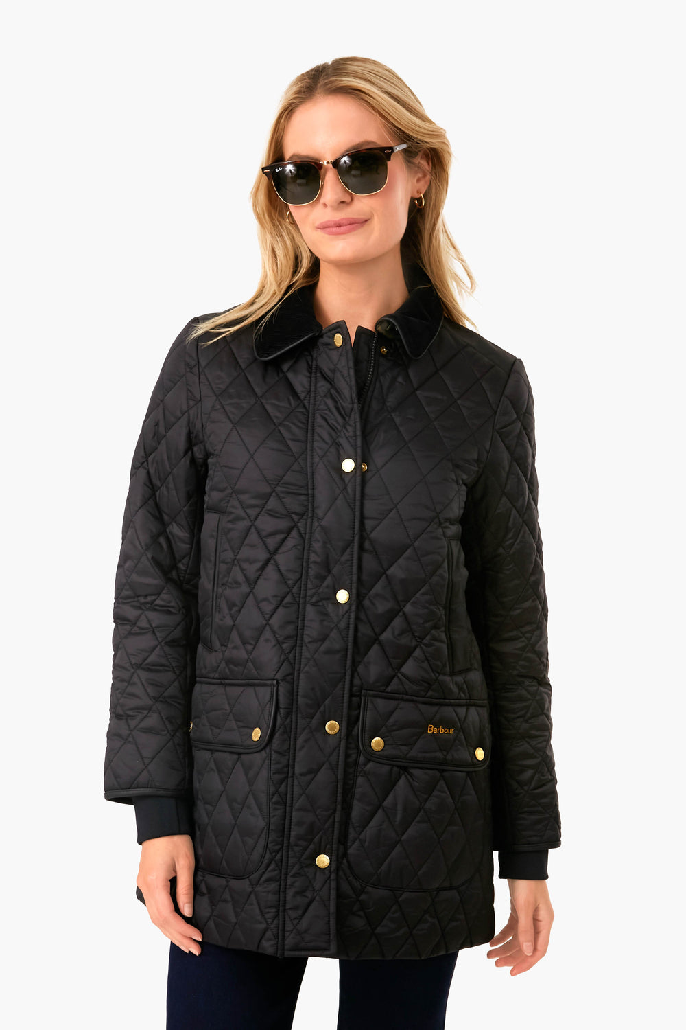 A woman wearing sunglasses and a Barbour quilted jacket with gold buttons stands facing forward.